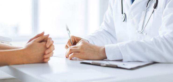 A doctor in a white coat with a stethoscope talks to a patient, holding a pen and clipboard, whilst the patient sits with hands folded on the desk.
