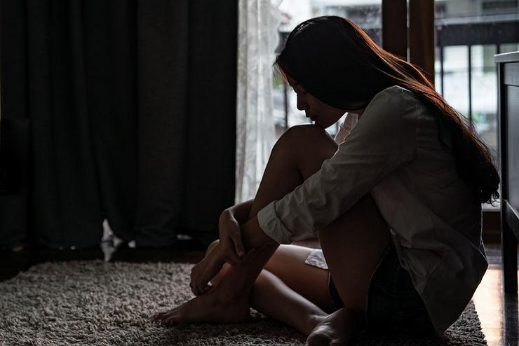 A person sitting on the floor in a dimly lit room, appearing deep in thought or contemplation. The scene conveys a sense of solitude and introspection, possibly related to anxiety or OCD. The soft lighting and shadows create a calm yet sombre atmosphere.