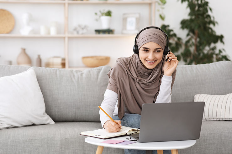 A woman wearing a hijab and headset sits on a sofa, smiling whilst taking notes in a notebook and looking at a laptop, engaged in online therapy in a cosy living room.