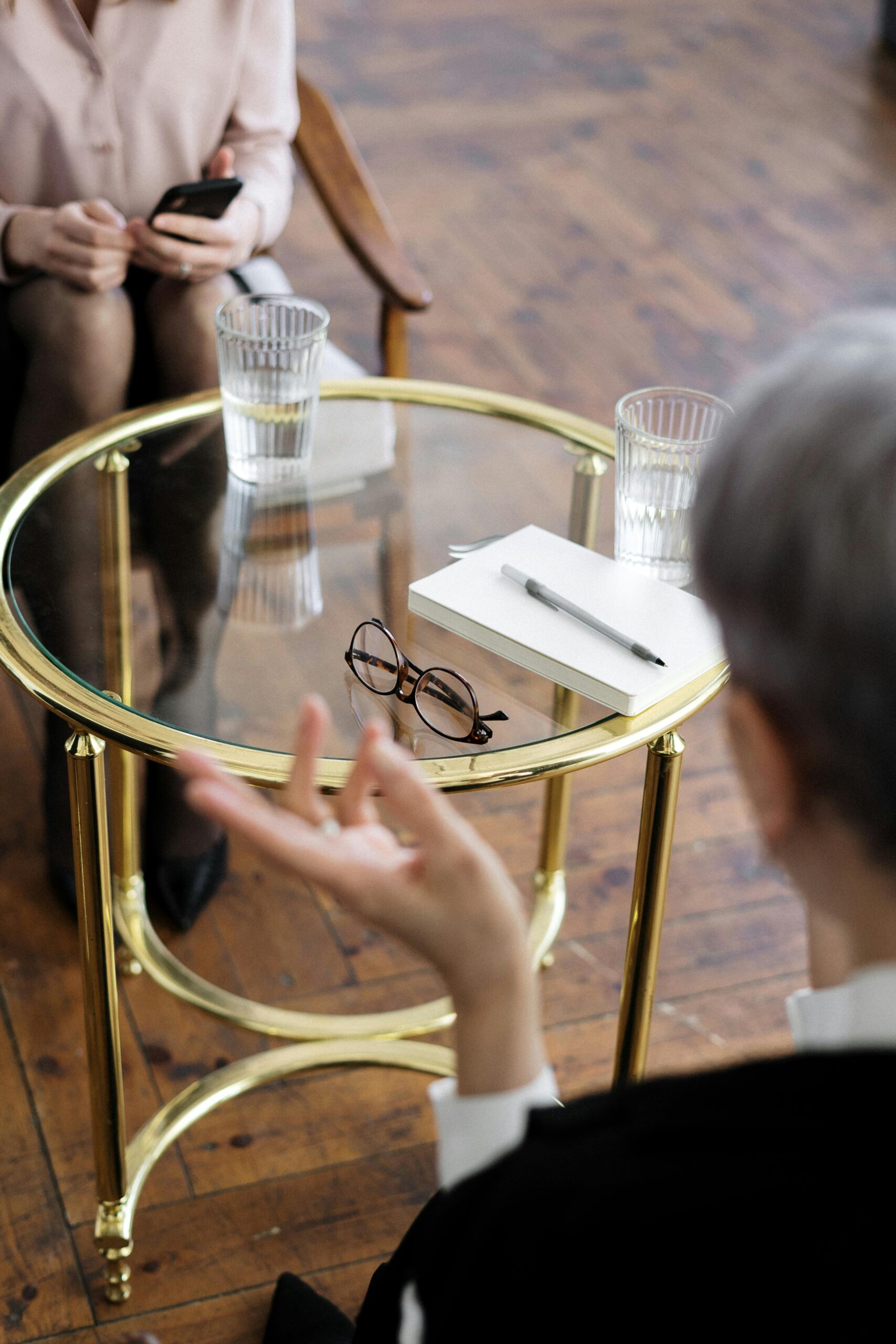 Two people sit in a comfortable, stylish setting engaged in a thoughtful conversation, suggesting a supportive mental health assessment or counseling session. A glass coffee table between them holds reading glasses, a notebook, a pen, and water glasses, while the warm wooden floor adds to the calm, welcoming atmosphere.